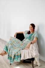 Meg in a green floral dress sitting on a blue velvet chair.
