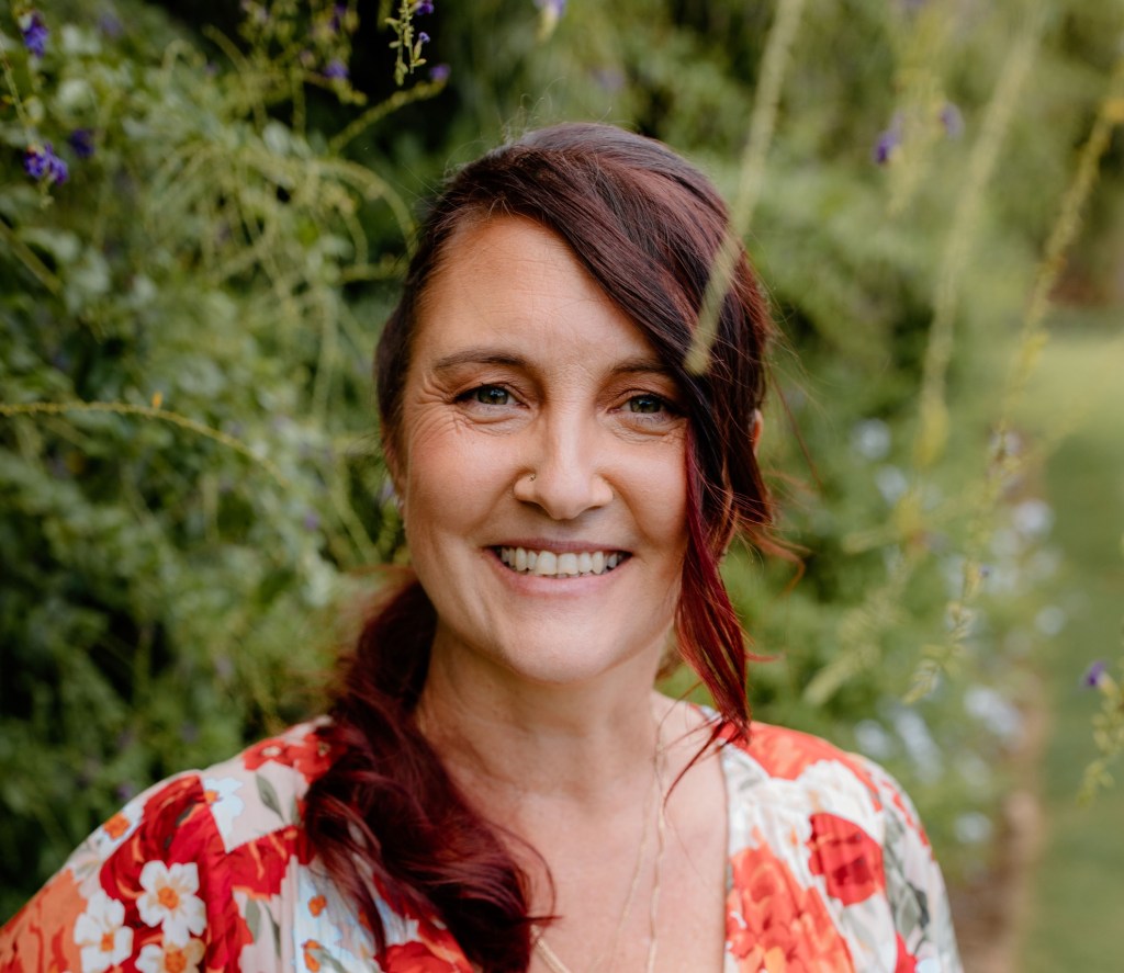 Picture of the author Meg standing in a garden. She wears a red floral dress and is smiling.